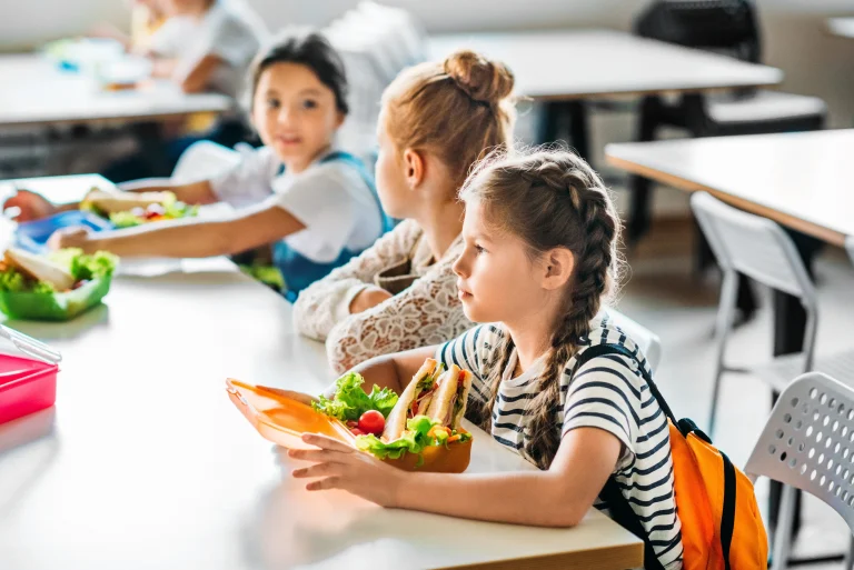 Dziewczynki w wieku wczesnoszkolnym jedzą drugie śniadanie z lunchboxów na szkolnej stołówce.