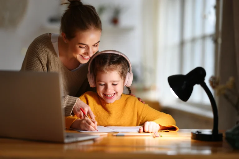 Mama pomaga córce w wieku szkolnym w nauce, pochylając się nad biurkiem i laptopem, przy których pracuje dziewczynka w żółtym swetrze ze słuchawkami na uszach.
